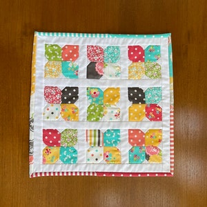 May include: A white quilted square with a patchwork design of colorful floral fabric. The fabric is arranged in a grid pattern with each square containing a four-petal flower design.
