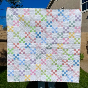 May include: A white quilt with a patchwork design featuring squares of various pastel colors, including pink, blue, green, and yellow. The squares are arranged in a random pattern, creating a visually appealing and intricate design.