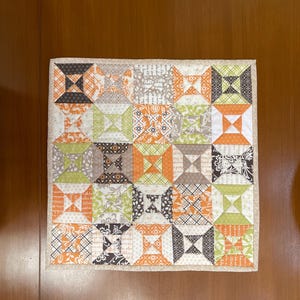 May include: A small quilted square with a patchwork design featuring various patterns in shades of orange, brown, green, and white. The quilt is framed with a beige border.