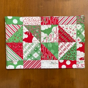 May include: A rectangular Christmas-themed quilted placemat. The patchwork design features red, green, and white fabrics with patterns including snowmen, candy canes, polka dots, and stripes. The edges are neatly finished with a coordinating border.