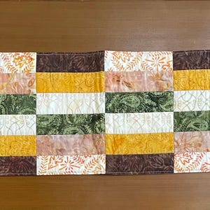 May include: A rectangular quilted table runner featuring a patchwork design of alternating fabric strips. The pattern includes shades of dark brown, mustard yellow, olive green, and cream, with various botanical and leaf-themed prints.