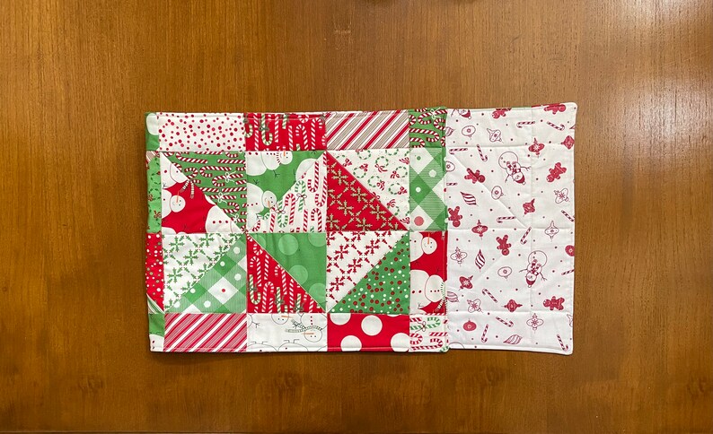 May include: A quilted Christmas-themed table runner. The patchwork design features red, green, and white fabrics with patterns of candy canes, snowmen, and polka dots. The runner is rectangular and appears to be handmade.