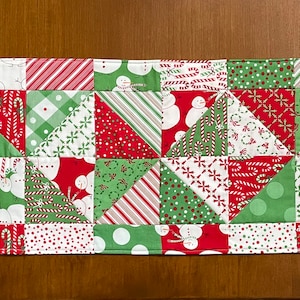 May include: A rectangular Christmas table runner with a patchwork design. The runner features red, green, and white fabrics with patterns including snowmen, candy canes, polka dots, and geometric shapes. The edges are neatly finished with a red border.