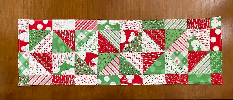 May include: A rectangular Christmas table runner with a patchwork design. The runner features red, green, and white fabrics with patterns including snowmen, candy canes, polka dots, and geometric shapes. The edges are neatly finished with a red border.