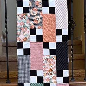 May include: A patchwork table runner with a variety of patterned fabrics, including floral, polka dot, and gingham designs. The runner features a black and white checkerboard pattern and is displayed on a wooden railing.