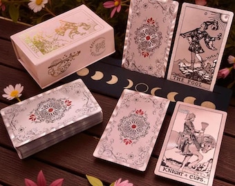 May include: A tarot card deck with a pink box featuring gold foil accents and the words "THE FOOL". The cards have intricate designs with red and silver details. The deck includes cards with the text "KNIGHT + CUPS".