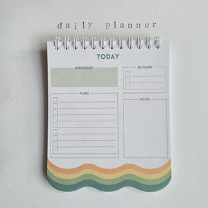 Könnte beinhalten: Ein spiralgebundener Tagesplaner mit dem Aufdruck "daily planner" oben. Der Planer hat Abschnitte für Aufgaben, Selbstpflege, Notizen und wichtige Dinge. Der untere Rand hat ein wellenförmiges Design in Grün-, Gelb- und Orangetönen.