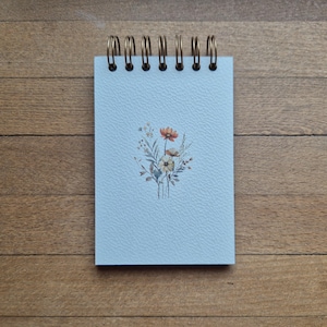 May include: A light blue spiral-bound notebook with a textured cover featuring a small, centred illustration of a wildflower bouquet in shades of orange, white, and green. The notebook is held together by a dark bronze-coloured metal wire coil.