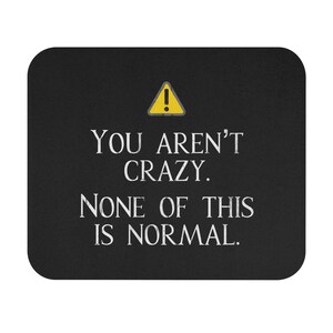 May include: Black rectangular mousepad with white text that reads "YOU AREN'T CRAZY. NONE OF THIS IS NORMAL." A yellow warning symbol is at the top. The mousepad is a desk accessory.