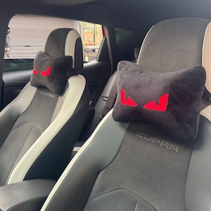 May include: Black headrest pillows with red eye-shaped designs are placed on the front seats of a car. The seats are gray and black with white accents. The word "CUPRA" is embroidered on the seat.