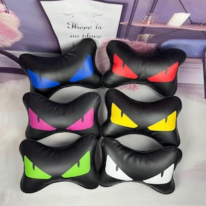 Könnte beinhalten: Eine Sammlung schwarzer, knochenförmiger Kissen, jedes mit einem Paar stilisierter Augen in verschiedenen Farben: Blau, Rot, Pink, Gelb, Grün und Weiß. Die Kissen sind auf einer hellen Oberfläche angeordnet.