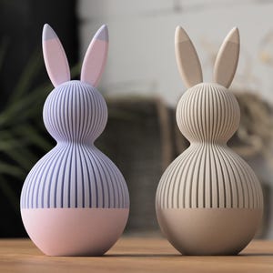 Könnte beinhalten: Zwei dekorative Hasenfiguren. Eine ist lavendelfarben mit rosa Akzenten, die andere beige. Beide haben ein geripptes Design und Hasenohren. Sie stehen auf einer Holzoberfläche.