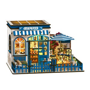May include: A miniature coffee shop model with a blue and white awning that reads "COFFEE". The model features a blue picket fence, a striped umbrella, and a small outdoor seating area. The shop is lit from within, showcasing interior details.