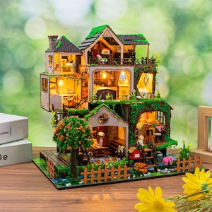 May include: A multi-story miniature dollhouse kit featuring detailed rooms with warm interior lighting, green moss-like roof accents, a small wooden fence, a tree structure, and various tiny furniture pieces arranged across several levels and balconies.