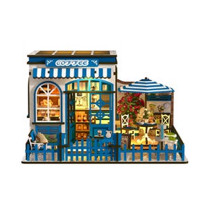 May include: Miniature coffee shop model with blue and white accents. Features a striped awning, a small outdoor seating area with a striped umbrella, and a decorative fence. The shop has a sign that says "Coffee".