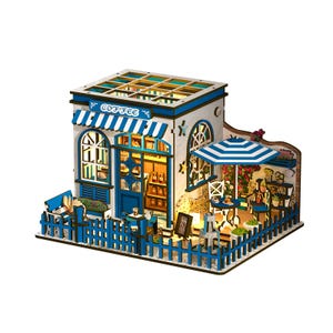 May include: A miniature wooden coffee shop model with a blue and white color scheme. The model features a striped awning, a small outdoor seating area with a blue and white umbrella, and a picket fence. The word "COFFEE" is visible on the awning.
