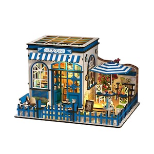 May include: A miniature coffee shop model with a blue and white color scheme. The model features a striped awning, a picket fence, and a small outdoor seating area with a striped umbrella. The word "COFFEE" is visible on the awning.