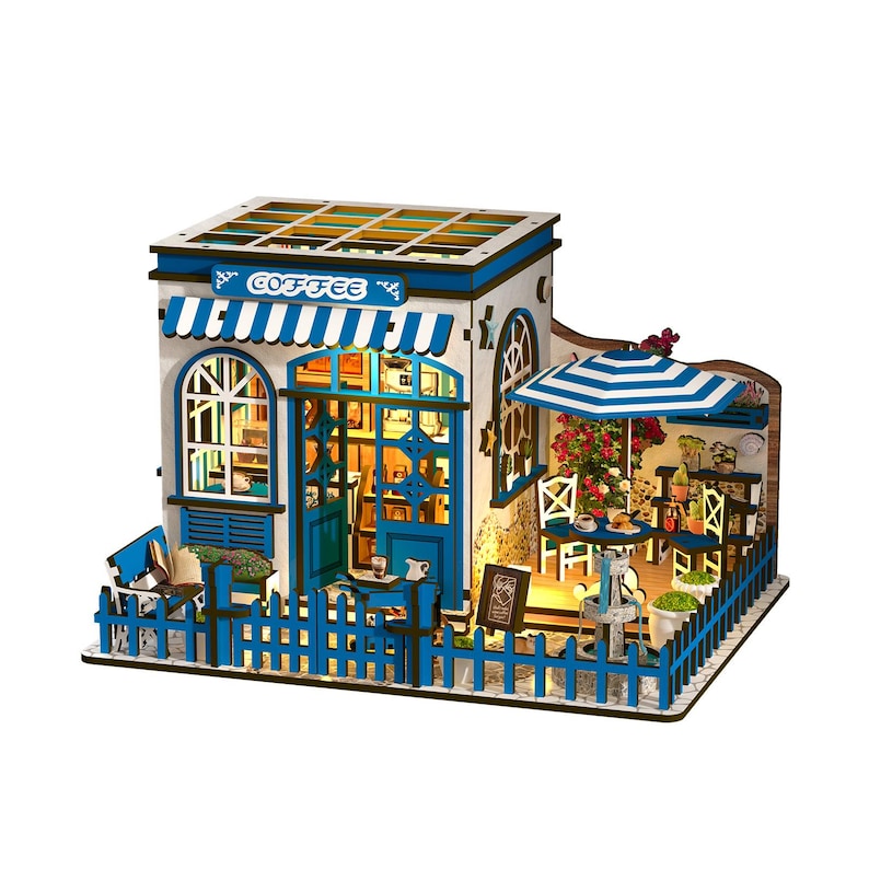 May include: A miniature coffee shop model with a blue and white color scheme. The model features a striped awning, a picket fence, and a small outdoor seating area with a striped umbrella. The word "COFFEE" is visible on the awning.