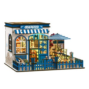 May include: Miniature coffee shop model with a blue and white color scheme. The model features a blue awning with the word "COFFEE", a blue picket fence, and a small outdoor seating area with a striped umbrella.