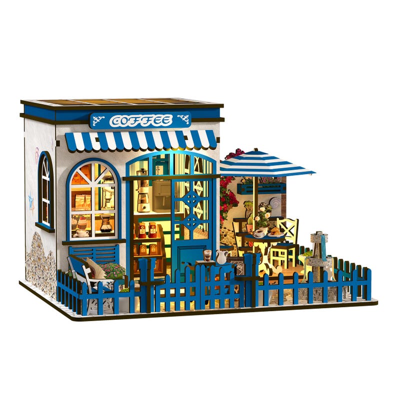 May include: Miniature coffee shop model with a blue and white color scheme. The model features a blue awning with the word "COFFEE", a blue picket fence, and a small outdoor seating area with a striped umbrella.