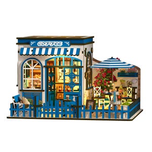 May include: A miniature coffee shop model with a blue and white striped awning and the word "COFFEE" above the entrance. The model features a blue picket fence, outdoor seating, and a small patio with a striped umbrella.