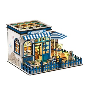 May include: A miniature wooden coffee shop model with a blue and white color scheme. The model features a striped awning, a small outdoor seating area with a table and chairs, and a decorative fence. The word "COFFEE" is displayed above the entrance.