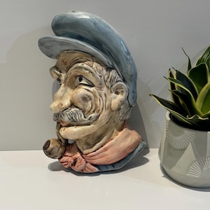 Peut inclure: Buste en céramique d'un vieil homme avec une casquette bleue, une moustache et une pipe. Le buste est peint dans des tons beige, rose et bleu. La tête de l'homme est légèrement tournée sur le côté.