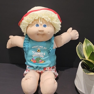 Peut inclure: Poupée Cabbage Patch Kid vêtue d'un haut bleu avec le texte "MEET ME at the FARMER'S MARKET" et d'un chapeau de paille à bord rouge. La poupée porte également un short à motifs de fraises. La poupée a les yeux bleus et les cheveux blonds.