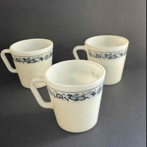 Puede incluir: Tres tazas de cerámica blanquecina con un diseño floral azul oscuro alrededor del borde. Cada taza tiene un asa y una forma ligeramente acampanada. Las tazas están dispuestas sobre una superficie gris oscuro.