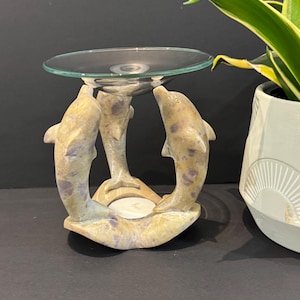 Peut inclure: Un bougeoir décoratif avec trois dauphins sculptés soutenant un plat en verre transparent. Les dauphins sont beiges avec des accents violets. Une bougie blanche est placée au centre de la base. Une plante d'intérieur dans un pot blanc est en arrière-plan.