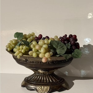 Peut inclure: Un bol décoratif sur piédestal de couleur bronze rempli de raisins artificiels. Les raisins sont un mélange de vert et de violet, avec des feuilles vertes. Le bol a un design orné et détaillé, pour la décoration intérieure.