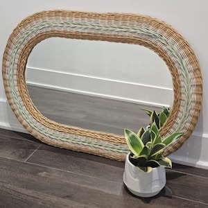 Könnte beinhalten: Ovaler Spiegel aus geflochtenem Rattan mit einem hellgrünen und naturfarbenen Rand. Der Spiegel lehnt an einer weißen Wand. Ein kleiner weißer Topf mit einer grün-gelben Pflanze steht davor.