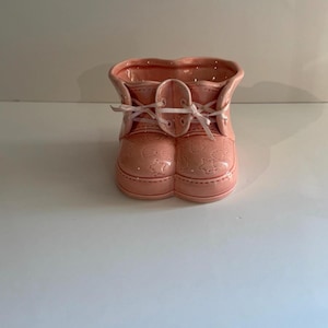 Op de afbeelding: Een roze keramische baby-laarsvormige plantenbak. De laars heeft een vetersluiting met witte lintveters en reliëfdetails van sterren en harten. De plantenbak heeft een zachte, pastelroze kleur.