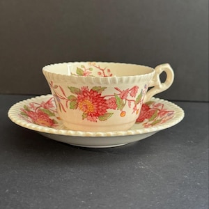May include: Cream-colored teacup and saucer set with a floral pattern in red, pink, and green. The cup has a handle and a scalloped rim, and the saucer has a matching design. The set is likely vintage or antique.