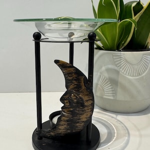 Peut inclure: Un brûle-parfum en métal noir avec un motif de croissant de lune à visage humain. Le support possède une base circulaire et soutient une coupelle en verre transparent sur le dessus. La lune présente une finition bronze et noire vieillie.