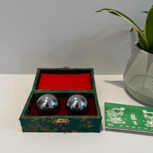 May include: Two shiny, silver metal health balls inside a green and red box with a gold clasp. The box has a floral pattern. A green paper with Chinese characters is next to the box. A potted plant is in the background.
