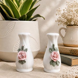 Peut inclure: Une paire de salières et poivrières en céramique blanche avec une texture côtelée verticale. Chaque pièce présente une rose rose sculptée en trois dimensions avec des feuilles vertes fixées au centre avant du corps blanc lisse et brillant.
