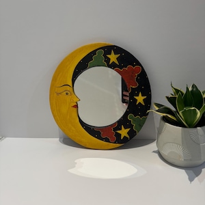 May include: A round mirror in the shape of a crescent moon, painted yellow with a smiling face. The black border features stars and colorful cloud shapes. A decorative piece for wall decor.