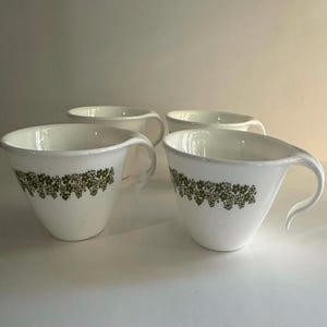 Peut inclure: Quatre tasses à thé en céramique blanche avec un motif floral vert sur le bord. Chaque tasse a une poignée unique. Les tasses sont disposées sur une surface blanche.