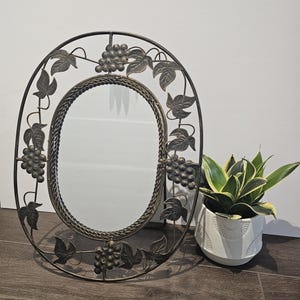 Peut inclure: Miroir ovale décoratif avec un cadre en métal bronze. Le cadre est orné de grappes de raisin et de feuilles. Un petit pot blanc à motifs géométriques avec une plante verte et jaune est à droite du miroir.