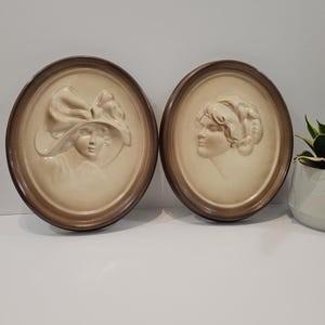 May include: Two oval wall plaques featuring cream-coloured relief portraits of faces. The left plaque shows a figure wearing a wide-brimmed hat with a large bow, while the right plaque depicts a profile view of a figure with styled hair. Both have brown frames.