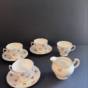 May include: A classic tea set comprising four teacups with saucers and a creamer. The white porcelain items are decorated with a pink rose design and gold edging. The set is displayed on a dark surface.