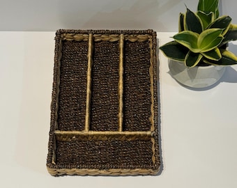 May include: A rectangular woven basket with four compartments, ideal for organizing utensils or flatware. The basket is made of dark brown and tan woven material. A small potted plant with green and yellow leaves sits to the right of the basket.