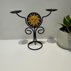 Puede incluir: Candelabro de metal negro con diseño de sol. El candelabro tiene dos brazos con plataformas circulares para velas. El diseño del sol está en el centro, con una cara amarilla y detalles en negro y naranja. La base tiene un diseño decorativo en espiral.