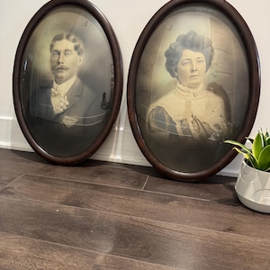 Puede incluir: Dos retratos ovalados antiguos en marcos de madera oscura. Los retratos son en blanco y negro, que representan a un hombre y una mujer. El hombre lleva un traje y la mujer un vestido. Los marcos se exhiben sobre un suelo de madera oscura.