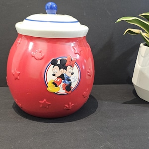 Peut inclure: Un pot à biscuits en céramique rouge orné de motifs en relief en forme d'étoiles et de têtes de Mickey Mouse. Le centre présente un graphique circulaire de Mickey et Minnie Mouse. Il comprend un couvercle blanc avec un bouton bleu.