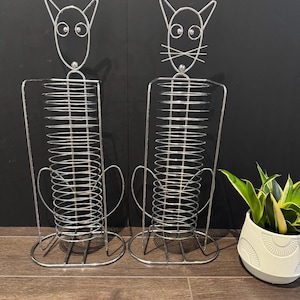 May include: Two cat-shaped wire metal organizers. Each has a cat face at the top with round eyes and pointed ears. The body is a series of horizontal wires. The base is a rounded wire frame. A potted plant is in the background.