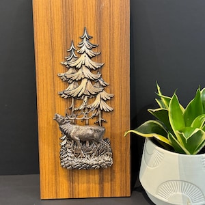 Puede incluir: Placa de pared rectangular de madera con un relieve metálico de un ciervo en un bosque. El ciervo está rugiendo, y los árboles están detallados con metal texturizado. La placa mide aproximadamente 30 cm de alto y 15 cm de ancho.