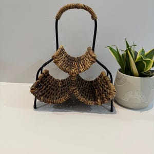 Peut inclure: Un porte-bouteille de vin décoratif avec un design de panier tressé. Le support est composé d'une armature en métal noir avec une poignée et de deux sections tressées pour maintenir la bouteille. Les sections tressées sont de couleur marron clair.