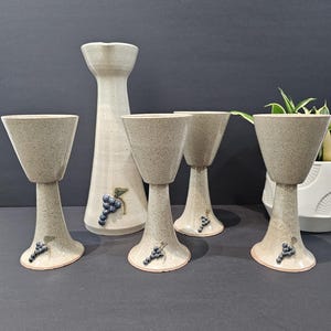 Peut inclure: Un ensemble en céramique composé d'un pichet haut et de quatre gobelets assortis. Chaque pièce présente un émail beige clair moucheté et est ornée d'un petit motif en relief représentant une grappe de raisin bleue près de la base.